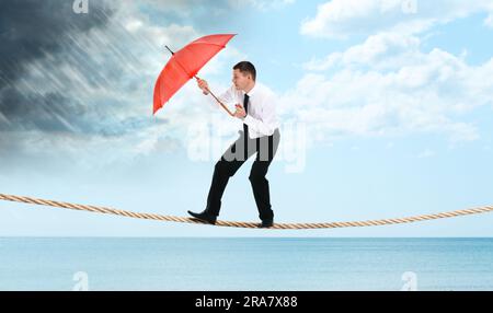 Risiken und Herausforderungen des Unternehmertums. Geschäftsmann mit Regenschirm, der vor kommenden Regenfällen am Seil über dem Meer schützt Stockfoto