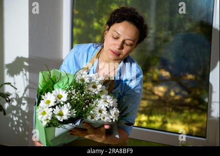 Eine bezaubernde, ethnische Frau mit Blumenmotiv und Dekorateurin hält einen wunderschönen Strauß aus weißen Gänseblümchen und Kamillenblumen in einem Studio mit Blumendesign für holi Stockfoto