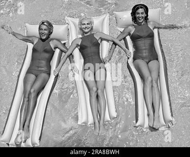 Vereinigte Staaten: ca. 1962 drei junge Frauen in zusammenpassenden Badeanzügen, die auf Flößen in einem Schwimmbecken zusammenschwimmen. Stockfoto