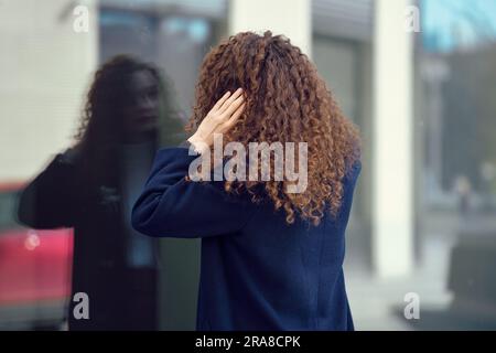 Eine unbekannte Frau im Mantel sieht ihr Spiegelbild im Fenster, während sie draußen ist Stockfoto
