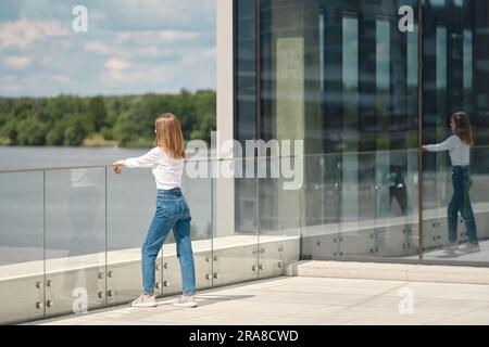 Eine nicht wiedererkennbare Frau steht auf der Aussichtsplattform, lehnt sich an ein gläsernes Geländer und schaut auf Wasser Stockfoto