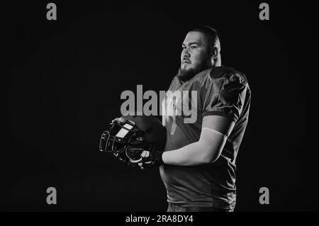 Ein asiatischer Mann mit rotem Bart in amerikanischer Fußballuniform trägt einen Helm auf schwarzem Hintergrund. Schwarzweiß. Stockfoto