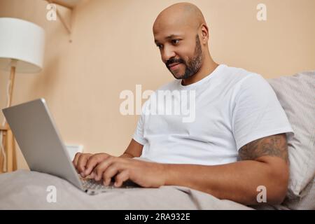 Porträt eines lächelnden schwarzen Mannes, der ein Notebook im Bett in einem gemütlichen Zuhause benutzt, Kopierbereich Stockfoto