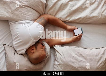 Minimale Draufsicht eines schwarzen Mannes, der im Bett liegt und ein Smartphone hält, mit weißer Bildschirmanzeige, Kopierbereich Stockfoto