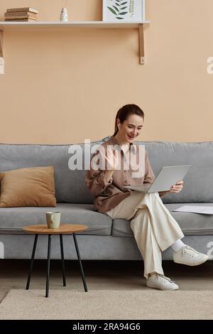 Vertikales, durchgehendes Porträt einer jungen, eleganten Geschäftsfrau, die von zu Hause aus arbeitet und auf dem bequemen Sofa zu einem Online-Meeting winkt Stockfoto