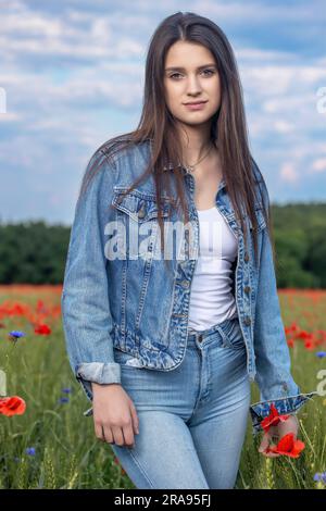 Das hübsche, langhaarige braune Mädchen in blauen Jeans steht auf einer Wiese mit rotem Mohn. Vertikal. Stockfoto