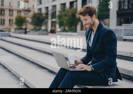 Zufriedener Geschäftsmann in blauem Anzug arbeitet außerhalb des Büros an einem Laptop und genießt eine sonnige Mittagspause. Stockfoto