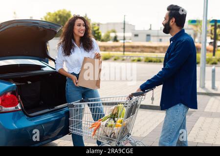 Glückliches arabisches Paar, das Einkaufstüten in einen Kofferraum auf einem Parkplatz packt, eine Dame, die mit ihrem Freund schaut und redet Stockfoto