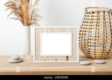 Minimalistisches Naturholzdekor mit weißem, leerem Bilderrahmen, trockenem Schilf in Vase und runder Holzlaterne mit Kerzenbrand. Hintergrundinformationen zu Innenräumen. Stockfoto