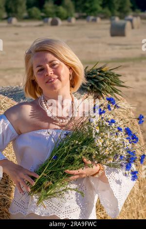 Eine ältere europäische Frau mit einem Blumenstrauß vor dem Hintergrund von Heuhaufen. Glücklicher Ruhestand in Europa. Stockfoto