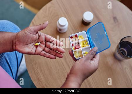 Handfläche einer älteren Frau, die eine kleine Kapsel und einen Behälter hält, mit Pillen über einen runden Holztisch mit zwei Plastikflaschen und einem Glas Wasser Stockfoto