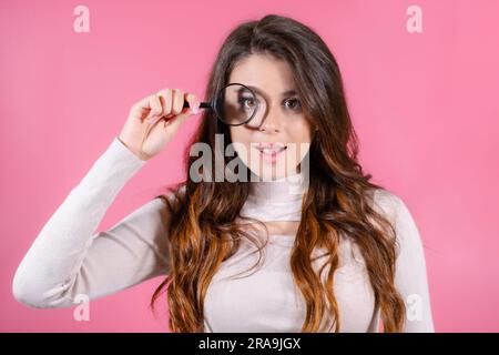 Porträt einer aufgeregten Frau, die steht, eine Lupe hält und auf den pinkfarbenen Hintergrund in die Kamera schaut Stockfoto