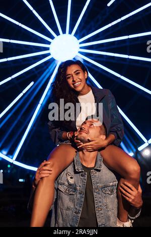 Junge Frau, die vor Lichtern von Riesenrad in die Kamera schaut. Ein Paar in der Nacht während des Festivals. Stockfoto