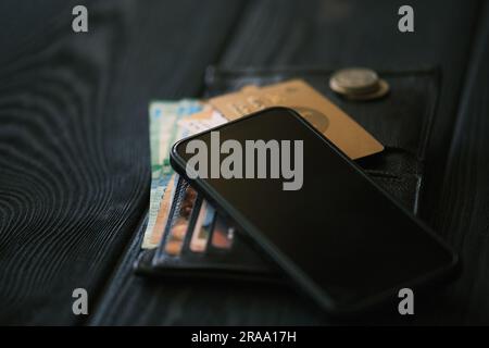 Ein Handy, das auf einer Ledertasche liegt, und ein kleiner Münzstapel auf einem schwarzen Holztisch, Finanzkonzept Stockfoto