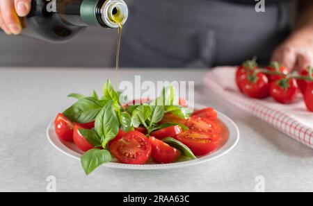 Ich gieße Olivenöl in eine Flasche über einen Kirschtomatensalat mit Basilikumblatt Stockfoto