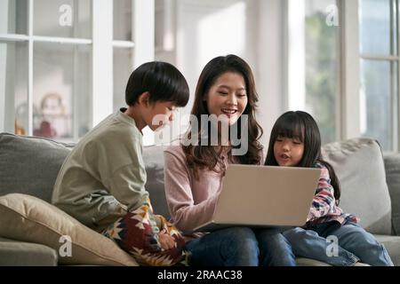 Junge asiatische Mutter und zwei Kinder, die zu Hause auf dem Familientisch sitzen und zusammen einen Laptop benutzen Stockfoto