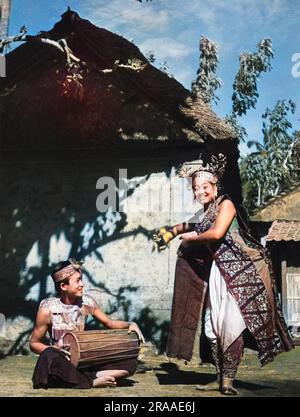 Bali, Indonesien : Eine Tänzerin beginnt ihre Routine als Hofnarr, gekleidet in farbenfroher und dekorierter Seide. Ein Schlagzeuger sitzt neben ihr. Datum: 1930er Stockfoto