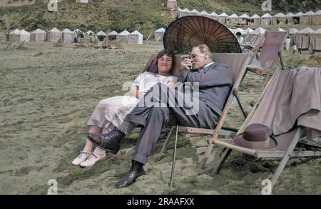 Ein Mann und eine Frau sitzen in Liegestühlen am Strand von Newquay, Cornwall, mit einem Sonnenschirm im orientalischen Stil, um sie vor der Sonne zu schützen. Datum: 1934 Stockfoto