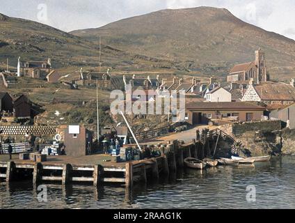 Eine Wasserszene in Schottland mit einem Steg, Häusern und einer Kirche. Fässer sind links aneinander gestapelt. Stockfoto