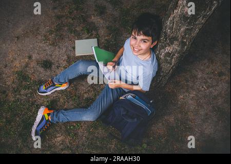 Blick von oben Teenager Schuljunge macht Hausaufgaben, sitzt nach der Schule unter einem Baum im Park und lächelt vor die Kamera Stockfoto