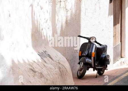 Ein schwarzer vespa-Motorroller parkt auf dem Bürgersteig in der Altstadt von Estepona, Spanien. Der Roller befindet sich neben einer alten, weiß gewaschenen Wand Stockfoto