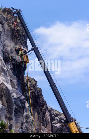 Ein Arbeiter verwendet einen Presslufthammer, um gefährliche überhängende Steine zu entfernen, während er an einem Käfig hängt, der an einem Kran hängt. Stockfoto