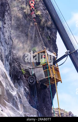 Ein Arbeiter verwendet einen Presslufthammer, um gefährliche überhängende Steine zu entfernen, während er an einem Käfig hängt, der an einem Kran hängt. Stockfoto