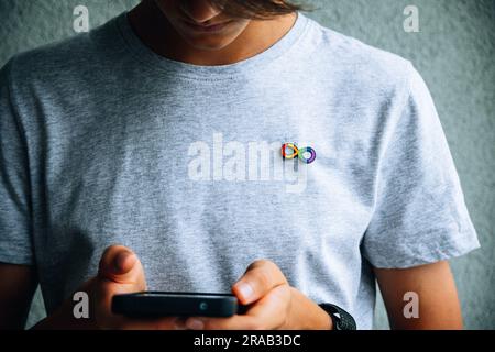 Teenager-Junge mit Autismus, unendlich, Regenbogen-Symbol, metallische Brosche auf dem T-Shirt mit Smartphone. Welttag des Autismus, Autismusrechte Stockfoto