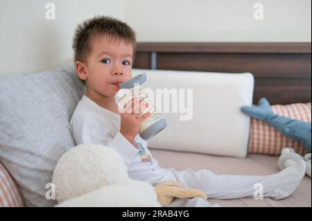 2 Jahre alter Junge sitzt auf dem Bett und trinkt fröhlich Milch aus einer mit Stroh ausgestatteten Flasche, umgeben von Spielzeugfreunden am Morgen in Pyjamas Stockfoto