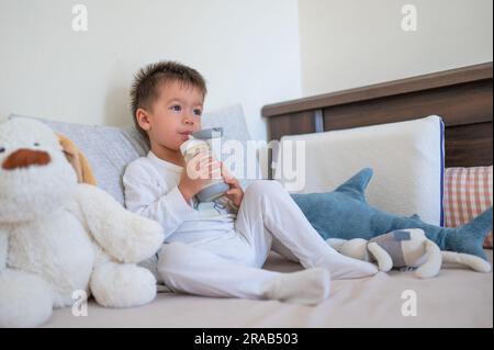 2 Jahre alter Junge sitzt auf dem Bett und trinkt fröhlich Milch aus einer mit Stroh ausgestatteten Flasche, umgeben von Spielzeugfreunden am Morgen in Pyjamas Stockfoto