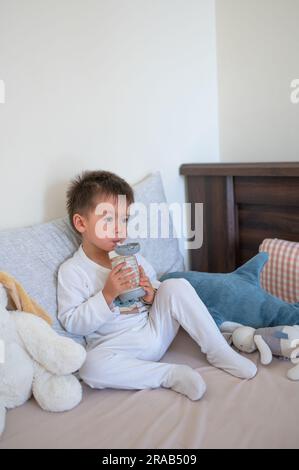 2 Jahre alter Junge sitzt auf dem Bett und trinkt fröhlich Milch aus einer mit Stroh ausgestatteten Flasche, umgeben von Spielzeugfreunden am Morgen in Pyjamas Stockfoto