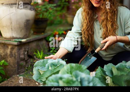 Junge Gärtnerin mit Gärtnerkelle im Strohhut, die sich um grüne Topfpflanzen kümmert – Nahaufnahme. Junge weiße Frau lächelt im Garten Stockfoto