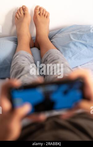 Ein Teenager im Schlafanzug am Morgen, der auf dem Bett liegt, spielt Videospiele, während seine Füße auf dem Kissen ruhen. Fokus auf Hintergrund und Kopierbereich. Stockfoto