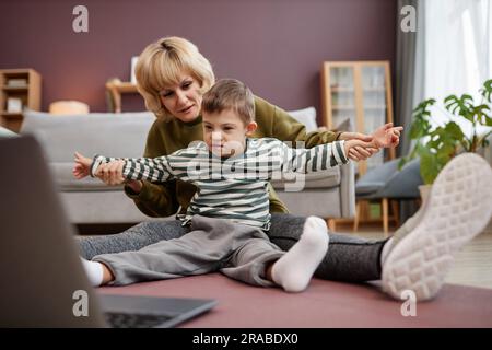 Vollständiges Porträt von Mutter und Sohn mit Daunensyndrom, während sie zusammen auf dem Boden zu Hause auf dem Laptop sitzen und Videos anschauen Stockfoto