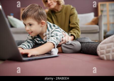 Nahaufnahme eines neugierigen, aufgeregten Jungen mit Down-Syndrom mit Laptop auf dem Boden zu Hause, Kopierraum Stockfoto