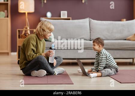 Seitliches Porträt einer Mutter und eines kleinen Jungen mit Down-Syndrom mit Laptop, während sie zu Hause auf dem Boden sitzen Stockfoto