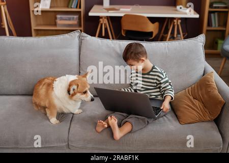 Hochwinkelporträt eines lächelnden kleinen Jungen mit Down-Syndrom, der ein Notebook benutzt, während er mit einem süßen Hund auf dem Sofa sitzt, Kopierraum Stockfoto