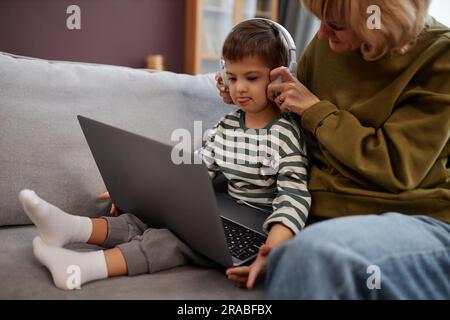 Porträt einer fürsorglichen Mutter, die dem kleinen Jungen mit Down-Syndrom Kopfhörer aufsetzt und sich online Videos ansieht, Kopierraum Stockfoto