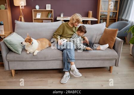 Ein langes Porträt von Mutter und Sohn mit Daunensyndrom, das zusammen Videos über einen Laptop ansieht, während er in einem gemütlichen Zuhause auf dem Sofa sitzt Stockfoto