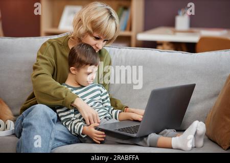 Seitliches Porträt von Mutter und Sohn mit Down-Syndrom, das den Laptop zusammen auf dem Sofa benutzt Stockfoto