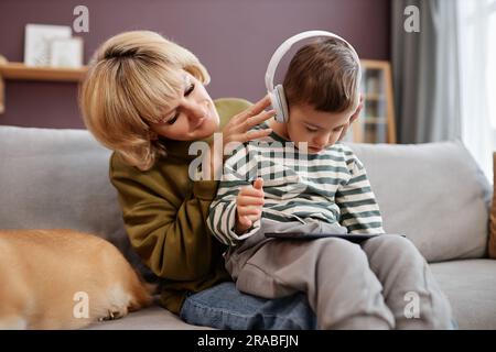 Liebevolle Mutter, die den kleinen Jungen mit Down-Syndrom mit Kopfhörern aufsetzt und zu Hause ein Notebook benutzt Stockfoto