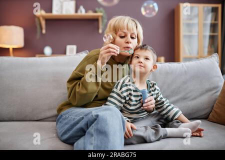 Porträt von Mutter und Kind mit Down-Syndrom, das beim Sitzen auf dem Sofa zu Hause Blasen bläst Stockfoto