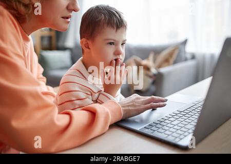 Nahaufnahme eines kleinen Jungen mit Down-Syndrom, der zu Hause studiert und mit Hilfe einer Mutter Computer benutzt Stockfoto