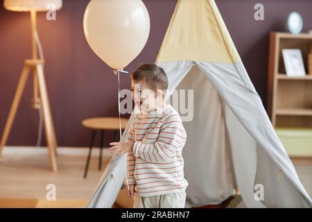 Porträt eines süßen kleinen Jungen mit Down-Syndrom, der Ballon hält, während er zu Hause spielt und lacht, Kopierraum Stockfoto