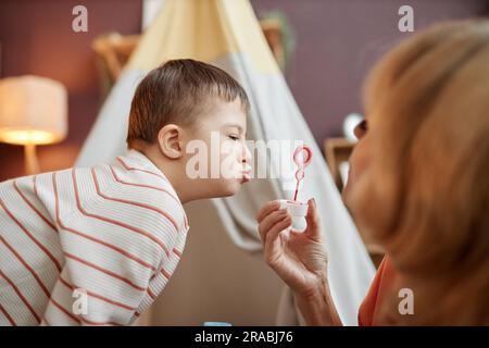 Porträt eines kleinen Jungen mit Down-Syndrom, der beim Spielen mit der Mutter im Haus Blasen bläst Stockfoto