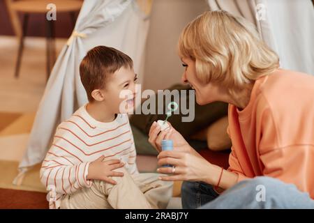 Porträt eines glücklichen kleinen Jungen mit Down-Syndrom, der beim Spielen mit der Mutter zu Hause Blasen bläst Stockfoto