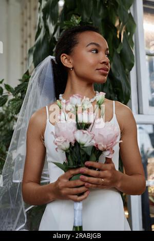 Vertikales Porträt einer schönen schwarzen Frau als junge Braut, die ein einfaches Hochzeitskleid trägt und einen Bouquet zur Seite hält Stockfoto