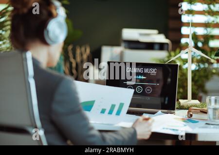 Moderne Frau mit Dokumenten und Laptop, die in einem modernen Büro mit Kopfhörern Musik hört. Stockfoto