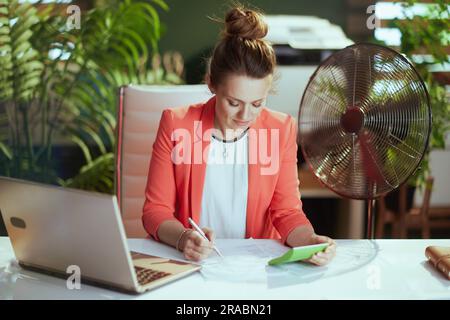 Nachhaltiger Arbeitsplatz. Moderne 40 Jahre alte Inhaberin eines kleinen Unternehmens in roter Jacke mit Taschenrechner, elektrischem Ventilator und Laptop. Stockfoto