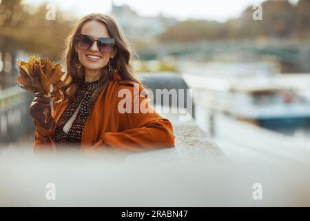 Hallo oktober. Fröhliche, trendige Frau im orangefarbenen Trenchcoat mit herbstgelben Blättern in der Stadt. Stockfoto